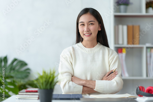 Beautiful Asian businesswoman sitting smiling, cross arms and opened her laptop and happily looking at the camera in the office.