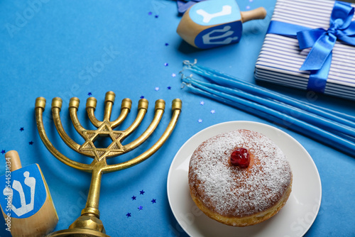 Plate with tasty doughnut, menorah, dreidels and candles for Hanukkah celebration on blue background photo
