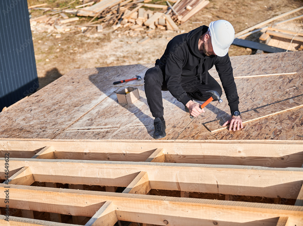 Carpenter hammering nail into OSB panel on the roof top of future cottage. Man worker building wooden frame house. Carpentry and construction concept. Carpenter hammering nail into OSB panel on the roof top of future cottage. Man worker building wooden frame house. Carpentry and construction concept.