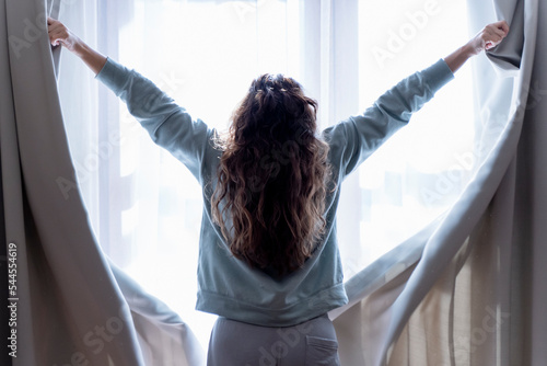 Happy young woman opens the curtains and looks out the window. Beautiful brunette opens the curtains to let the morning sunlight