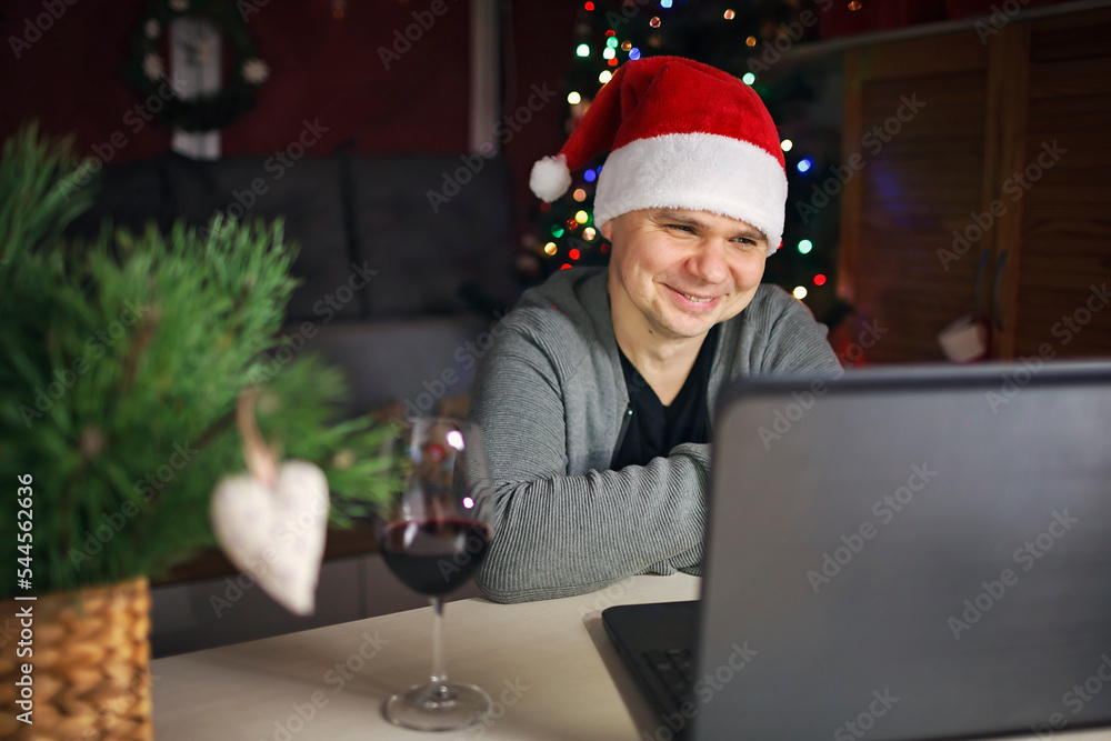 Online Christmas celebration. Happy man in Santa red hat celebrating with family virtually via internet and notebook. Video call. Refugees and immigrants, distant holiday, lifestyle Online Christmas celebration. Happy man in Santa red hat celebrating with family virtually via internet and notebook. Video call. Refugees and immigrants, distant holiday, lifestyle