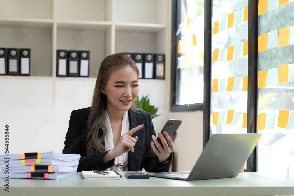 Smiling beautiful Asian businesswoman analyzing chart and graph showing changes on the market and holding smartphone at office. Smiling beautiful Asian businesswoman analyzing chart and graph showing changes on the market and holding smartphone at office.