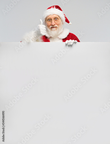 Handsome Santa Claus in glasses and white gloves holds a large vertical blank board and looks at the camera. Big white banner with Santa Claus showing thumbs up. © orientka