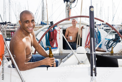 man topless with a bottle of beer in his hand sits on the deck of a yacht in seaport