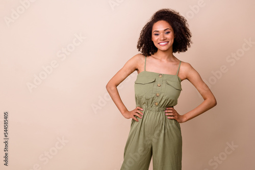 Photo of gorgeous cute nice satisfied woman with wavy hairstyle dressed khaki overall arms on waist isolated on beige color background