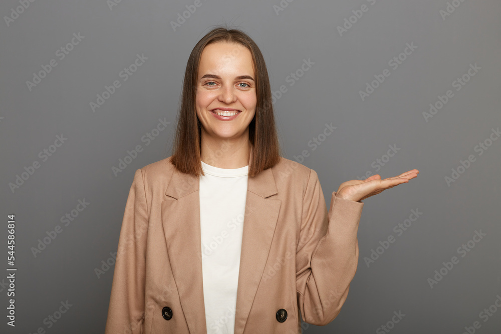 Portrait of satisfied positive happy woman with dark hair wearing jacket presenting copy space on palm, showing empty place for commercial text or goods, standing against gray wall. Portrait of satisfied positive happy woman with dark hair wearing jacket presenting copy space on palm, showing empty place for commercial text or goods, standing against gray wall.