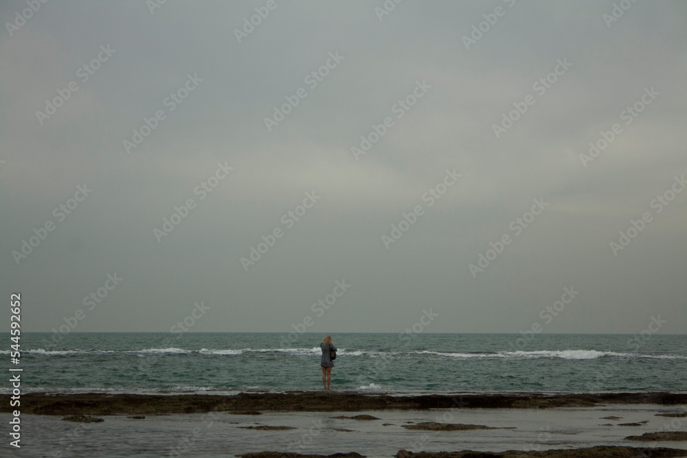 blonde woman in a short dress walking above the sea looking at her reflection in the water on a cloudy day in the bay of Cadiz.