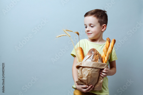 child with a grocery bag with freshly baked bread and ears of wheat, smiling softly against a uniform background.