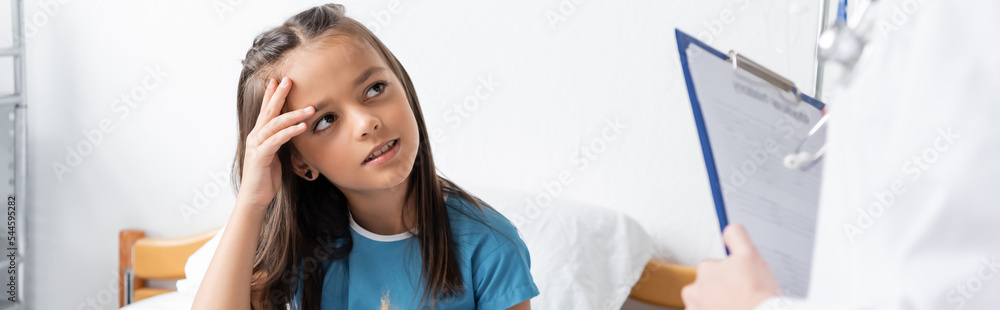 Child touching head near blurred doctor with clipboard in hospital, banner. Child touching head near blurred doctor with clipboard in hospital, banner.