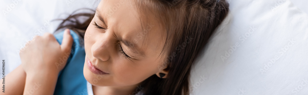 High angle view of ill child touching shoulder while lying on bed in hospital ward, banner. High angle view of ill child touching shoulder while lying on bed in hospital ward, banner.