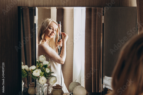 Woman curls her hair on curling iron in front of mirror at dressing table. Preparing for party photo