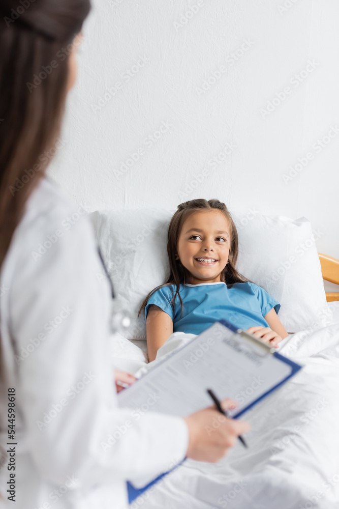 Smiling kid lying on bed while blurred doctor holding clipboard in hospital ward. Smiling kid lying on bed while blurred doctor holding clipboard in hospital ward.
