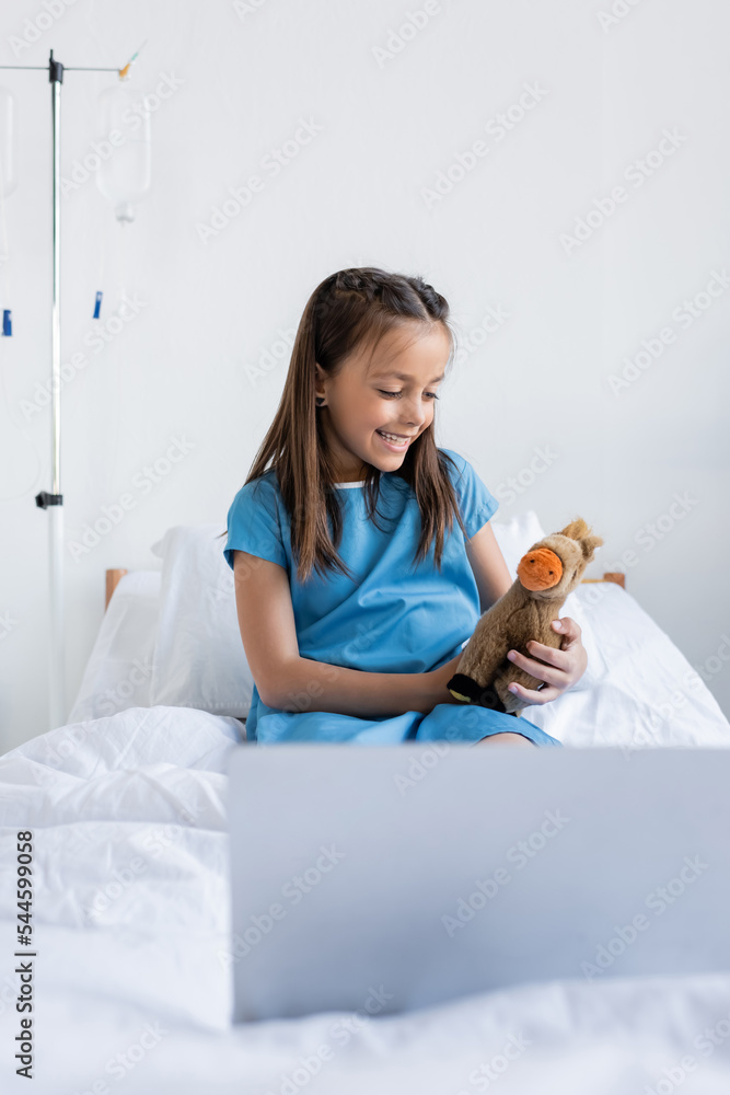 Cheerful kid holding toy near blurred laptop on hospital bed. Cheerful kid holding toy near blurred laptop on hospital bed.