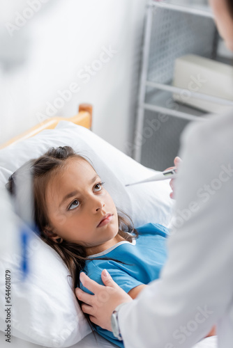 Doctor holding thermometer and touching child on clinic bed.