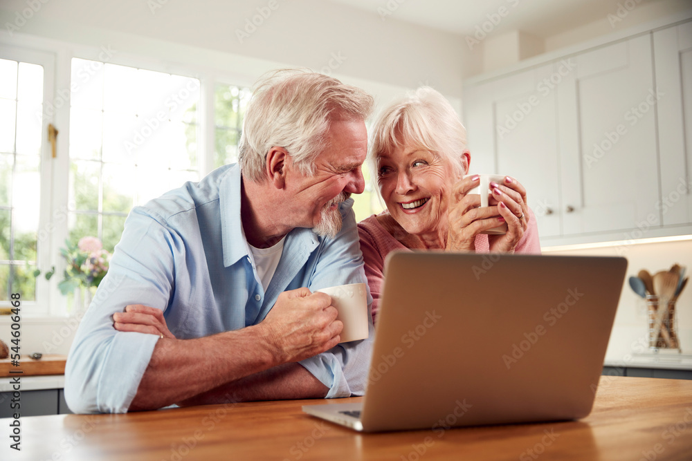Retired Senior Couple Sitting In Kitchen At Home Drinking Coffee And Using Laptop