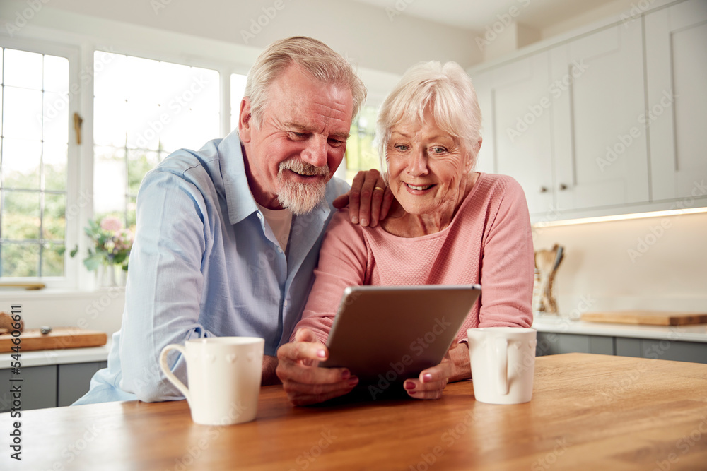 Retired Senior Couple Sitting In Kitchen At Home Drinking Coffee And Using Digital Tablet