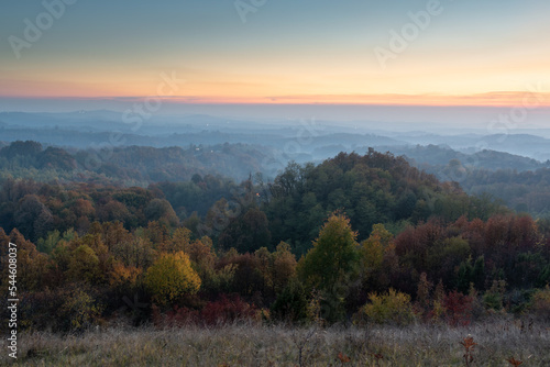 Rural landscape during dusk in autumn, hilly countryside with lush colourful forests and orange glow in sky