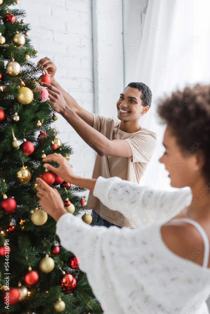 happy african american man decorating christmas tree near girlfriend on blurred foreground happy african american man decorating christmas tree near girlfriend on blurred foreground