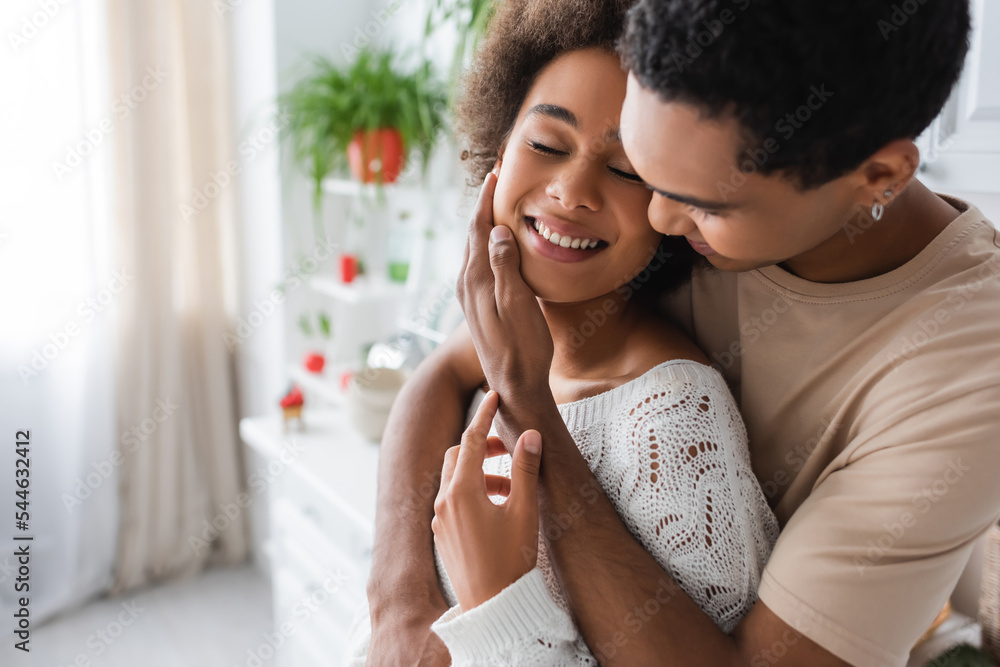 young african american man hugging and touching face of happy woman with closed eyes in kitchen young african american man hugging and touching face of happy woman with closed eyes in kitchen
