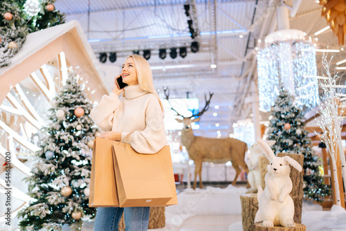 Low-angle view of pretty blonde young woman talking smartphone and holding paper bags with purchase standing in hall of celebrate shopping mall in Christmas eve, on background of xmas decorations.