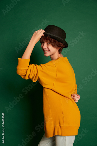 Portrait of cheerful teen girl with curly brown short hair posing in yellow sweater and hat isolated over green background
