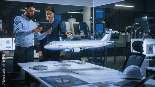 Aeronautics Factory Office Meeting Room: Chief Engineer Holds Tablet Computer, Showing Augmented Reality Airplane to a Female Project Manager, They Test Aerodynamics. Modern Industry 4.0 Research. photo
