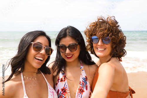 Three beautiful women taking a selfie on the beach with the sea in the background. The women are on holiday and are smiling for the photo, wearing sunglasses. Holiday and travel concept.
