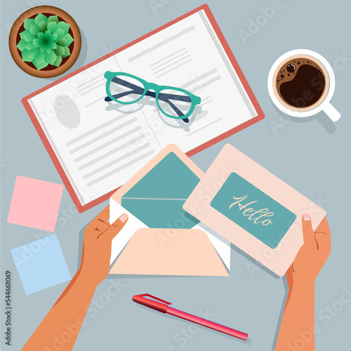 Hands on the table tear off an envelope with a postcard. Top view. Notepad, pen, glasses, coffee cup, flower.