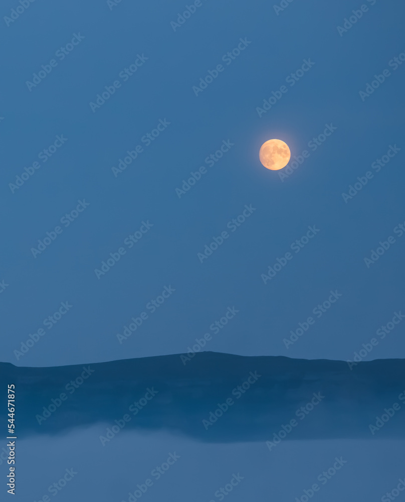 Full moon in the mountains in the early morning with a mountain cliff and dense clouds in the valley, orange moon in the autumn morning in the mountains Full moon in the mountains in the early morning with a mountain cliff and dense clouds in the valley, orange moon in the autumn morning in the mountains