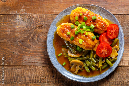 Chicken cutlets with green beans and mushrooms in tomato sauce in a plate next to the table with garlic and a knife.copy space