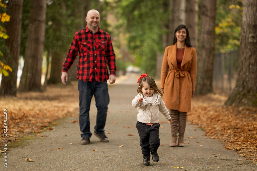 A cute little girl is playfully running away from her parents in a beautiful autumn colored park