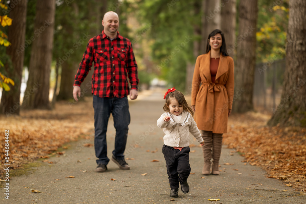 A cute little girl is playfully running away from her parents in a beautiful autumn colored park A cute little girl is playfully running away from her parents in a beautiful autumn colored park