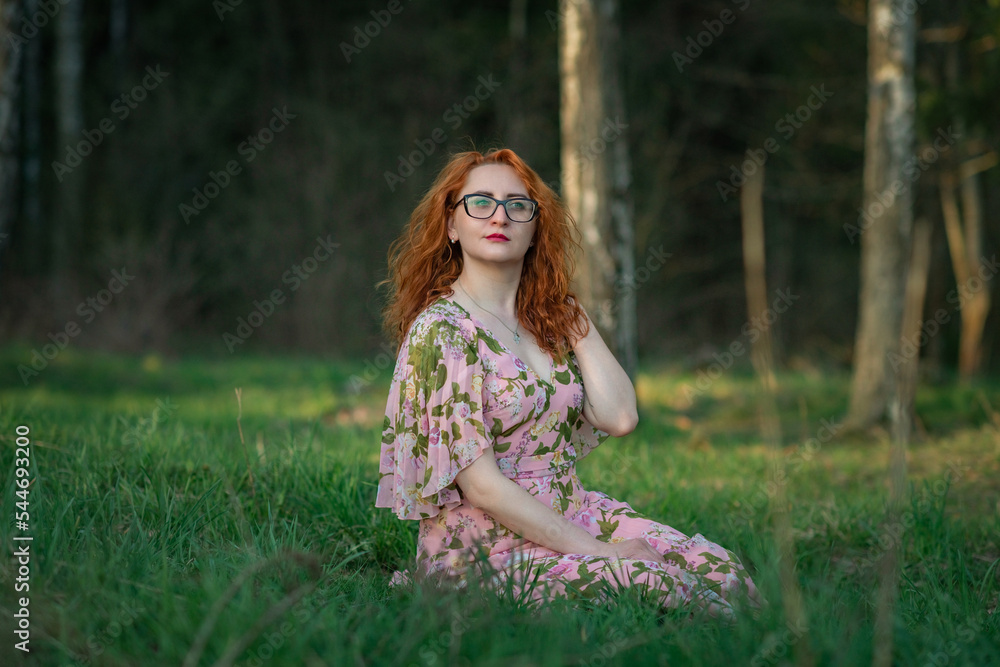Portrait of a young beautiful red-haired girl in a dress sitting in the forest on the grass.