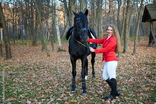 Young sexy woman standing near a horse in a red suit. Beautiful professional female jockey standing near horse. A cute female in a red jacket and white pants. The girl caresses her favorite horse. 