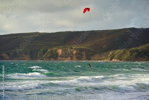 Kiteboarder jumping with red kite on the water. Water sportsman with twintip board between waves in front of mountain landscape. France, Normandy, Manche, Vauville.