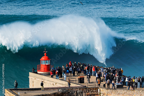 People watching the big giant waves crashing near the Fort of Nazare Lighthouse in Nazare, Portugal. Biggest waves in the world. Touristic destination for surfing and lovers of radical sports.
 photo