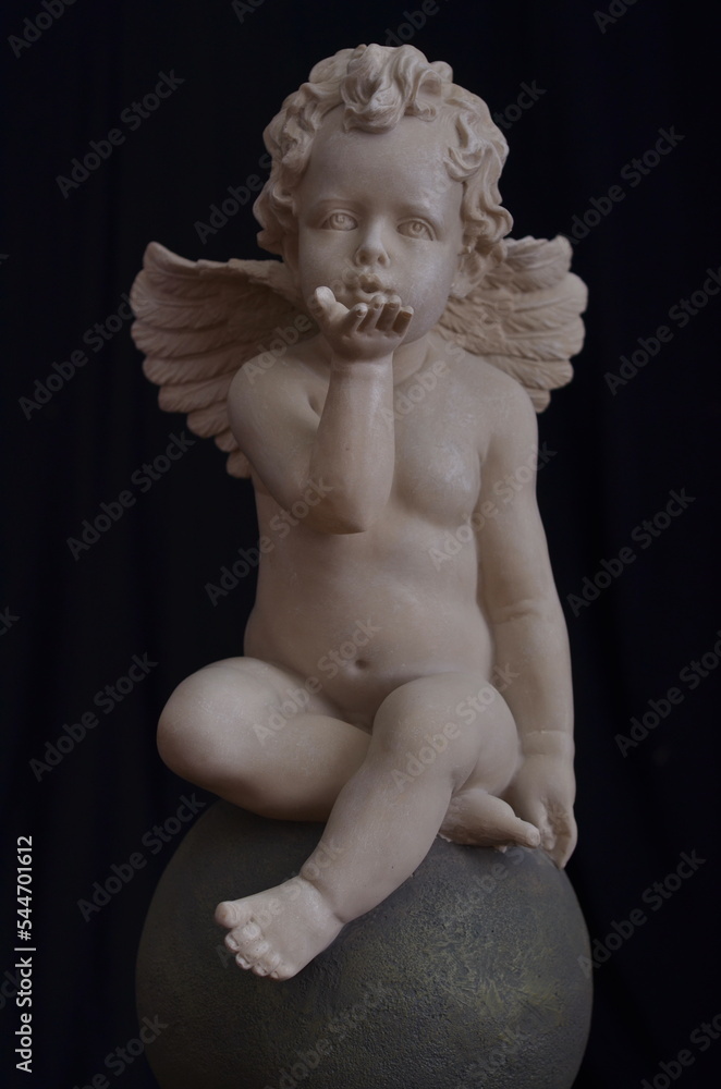 A marble angel with wings sits on a round stone and blows a kiss A marble angel with wings sits on a round stone and blows a kiss