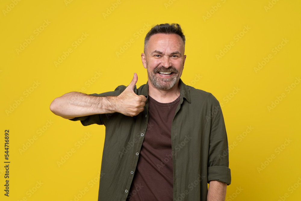 Adult mature beard man over isolated yellow background doing happy thumbs up gesture with hand. Approving expression looking at camera with showing success. Adult mature beard man over isolated yellow background doing happy thumbs up gesture with hand. Approving expression looking at camera with showing success.