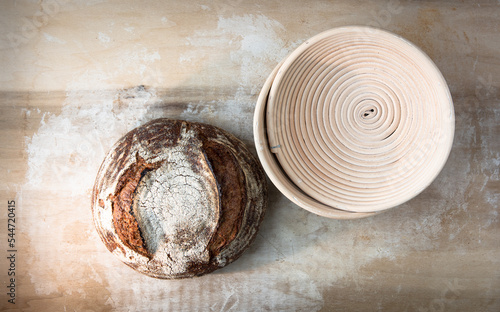 Single round loaf of crusty baked sourdough bread on lightly floured blonde wood board with coiled proofing basket.