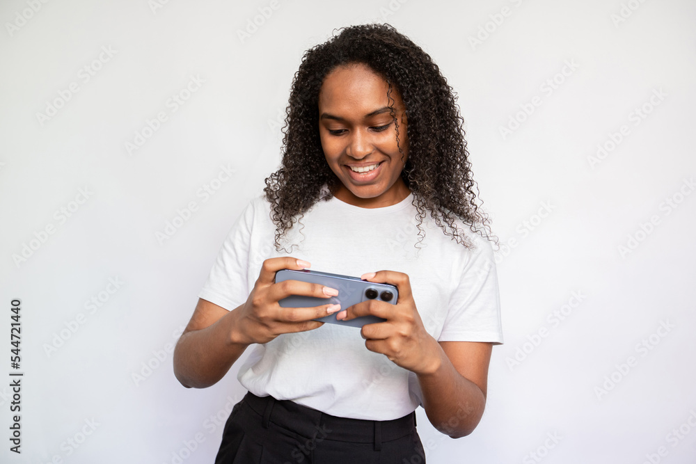 Portrait of happy young woman watching video on mobile phone. African American woman wearing white T-shirt playing games on smartphone. Mobile technology concept Portrait of happy young woman watching video on mobile phone. African American woman wearing white T-shirt playing games on smartphone. Mobile technology concept