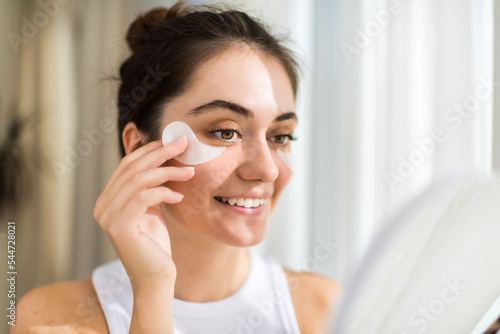A young caucasian smiling woman with patches under her eyes looking in the mirror. Skin care theme