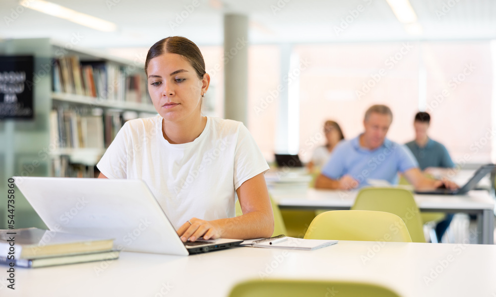 Fototapeta premium Portrait of young woman studying at library using books and laptop