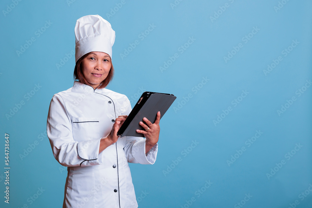 Asian cook with apron and white uniform holding tablet computer browsing dish recipe on gastronomy site. Cheerful senior chef cooking culinary dinner using healthy ingredient, food preparation concept Asian cook with apron and white uniform holding tablet computer browsing dish recipe on gastronomy site. Cheerful senior chef cooking culinary dinner using healthy ingredient, food preparation concept