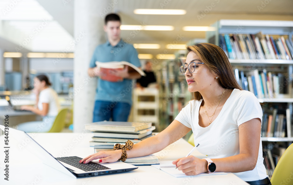 Fototapeta premium Positive young lady in casual clothes and glasses working remotely using a computer in a quiet library