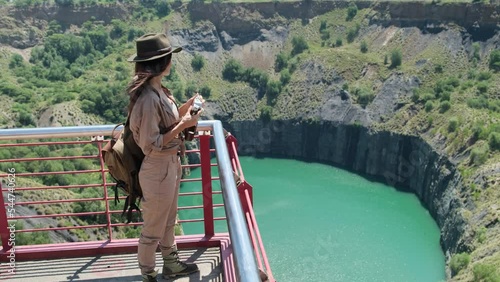 woman traveler in a hat and a backpack makes a photo near a kimberlite pipe. Diamond quarry open pit top down view, on sunny summer day. Mining equipment for the transportation of minerals. photo