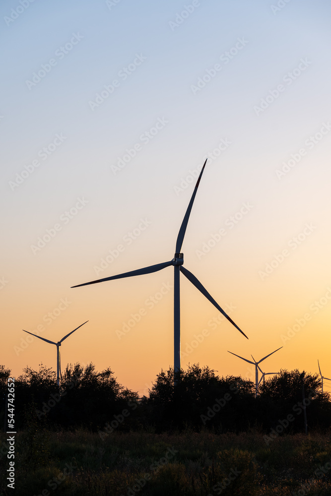 Large windmill against backdrop of rising sun in yellow and blue tones behind small green forest. Large windmill against backdrop of rising sun in yellow and blue tones behind small green forest.
