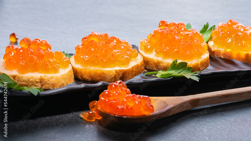 Red caviar in wooden spoon and on butter buns on black plate with green parsley on sides. Red luxury caviar on wooden spoon and on butter rolls on gray or black background. Red caviar in wooden spoon and on butter buns on black plate with green parsley on sides. Red luxury caviar on wooden spoon and on butter rolls on gray or black background.