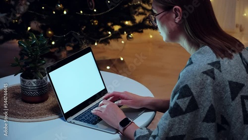 Blank white screen laptop mockup, woman using laptop typing on the keyboard at home on background of christmas tree and lights