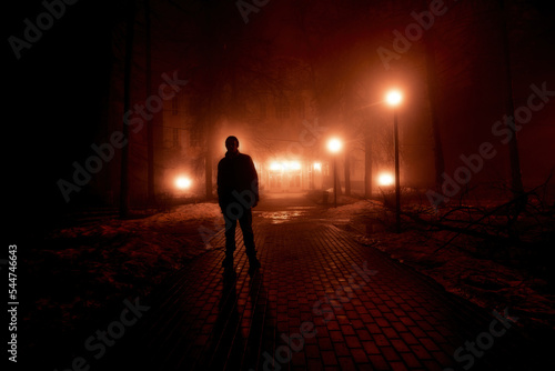 Sad man alone walking along the alley in night foggy park