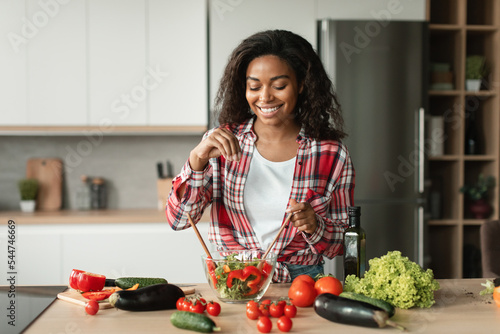 Homemade food and health care. Cheerful black young woman chef in casual salts salad from organic vegetables photo